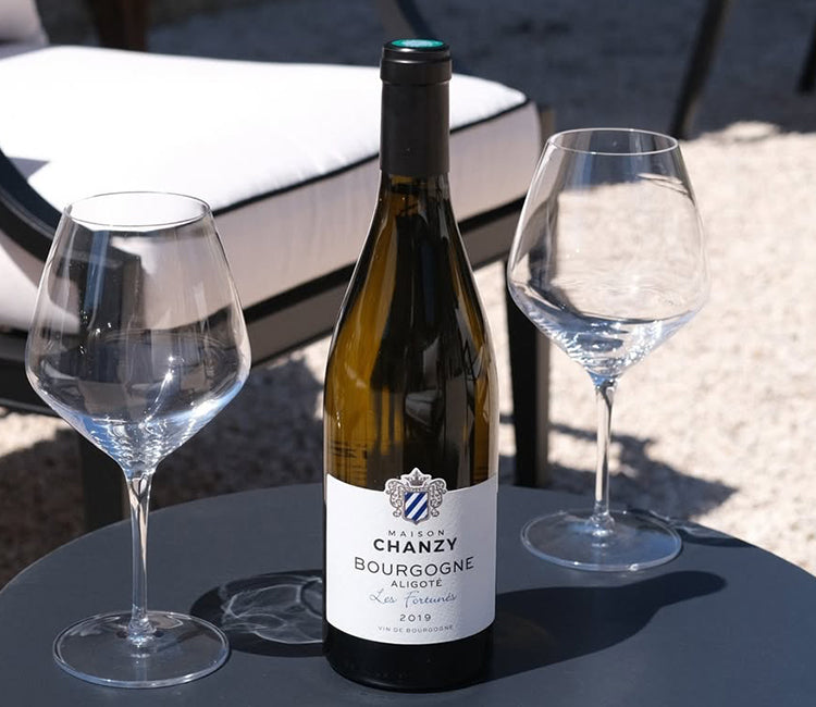 Maison Chanzy, Bourgogne Aligoté, hvidvin fra Bourgogne - Flaske Maison Chanzy Bourgogne Aligoté 2019 med to vinglas på bord i solen.